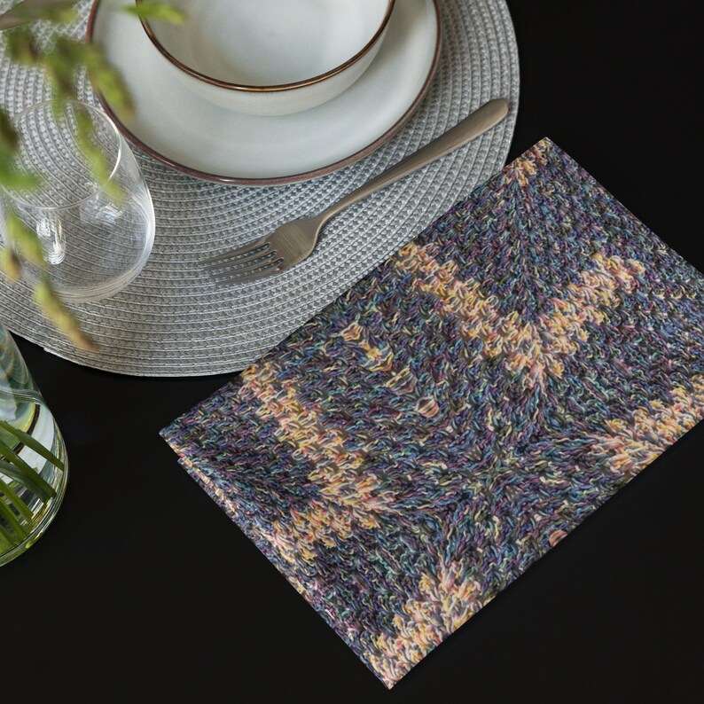 Puede incluir: Una servilleta de tela plegada, estampada y multicolor, est&aacute; sobre una superficie negra. Tambi&eacute;n se ven un cuenco y un plato blancos con borde marr&oacute;n, un tenedor plateado y un vaso transparente. La servilleta tiene un aspecto tejido y texturizado.