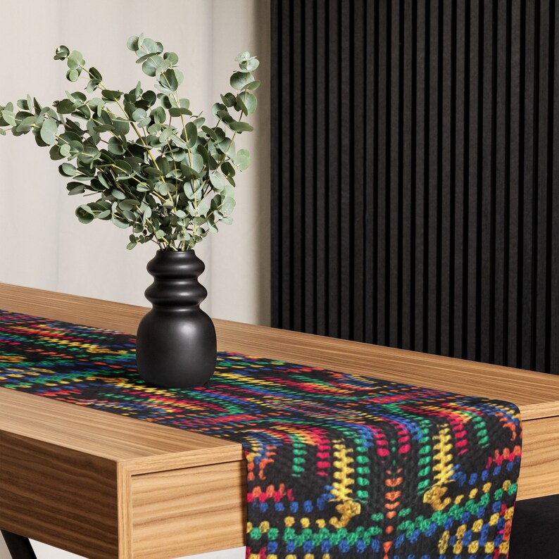 May include: A black table runner with a colorful, geometric pattern of red, yellow, green, and blue. A black vase with eucalyptus branches sits on a wooden table. The runner adds a vibrant touch to the table setting.