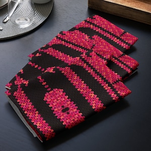 May include: Three black cloth napkins featuring a repeating pattern of vertical stripes and floral-like designs in pink and red hues. The napkins are folded and stacked, suggesting a dining or kitchen setting. The napkins are on a dark surface.