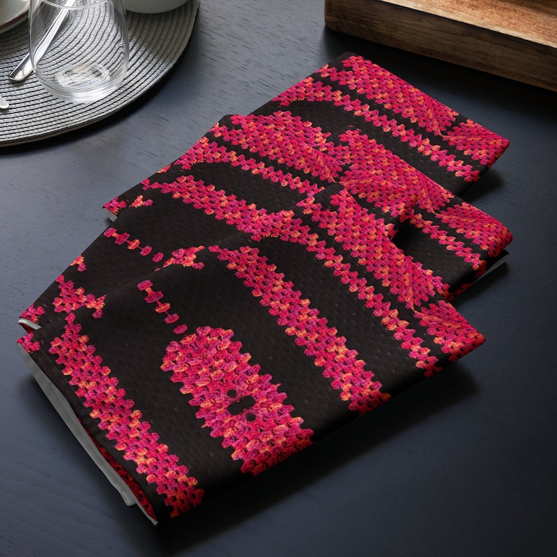 May include: Three black cloth napkins featuring a repeating pattern of vertical stripes and floral-like designs in pink and red hues. The napkins are folded and stacked, suggesting a dining or kitchen setting. The napkins are on a dark surface.