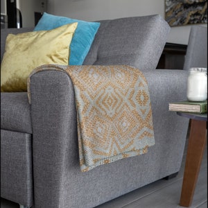 Pode incluir: Um sof&aacute; cinza com uma manta estampada em bege e branco, drapeada sobre o bra&ccedil;o. Duas almofadas, uma amarela e outra azul, est&atilde;o no sof&aacute;. Uma mesa lateral com uma vela e um livro.