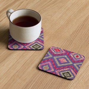May include: Two square coasters with a geometric pattern in shades of purple, pink, red, and black. One coaster has a white mug with tea on top. The coasters are on a light wood surface.