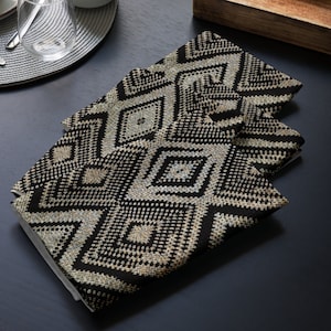 May include: Four folded black and beige geometric patterned cloth napkins. The napkins feature a repeating diamond design. The napkins are made of a textured fabric and are stacked on top of each other.