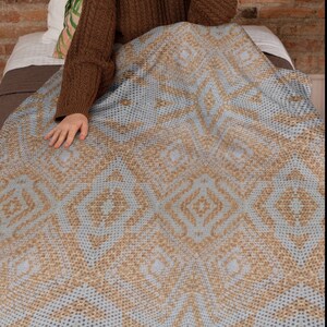 Pode incluir: Um cobertor azul claro e bege com um padr&atilde;o geom&eacute;trico. O cobertor est&aacute; sobre uma cama, com o bra&ccedil;o e a m&atilde;o de uma pessoa vis&iacute;veis. O padr&atilde;o do cobertor apresenta formas de diamante.