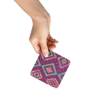 May include: A square coaster with a geometric pattern held by a hand. The coaster features a repeating diamond design in shades of purple, red, pink, and yellow. The coaster is made of a woven material.