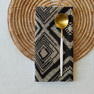 May include: A gold-colored spoon with a white handle rests on a black and beige patterned napkin. The napkin is placed on a round, woven placemat. The setting is on a light gray surface, creating a neutral dining scene.
