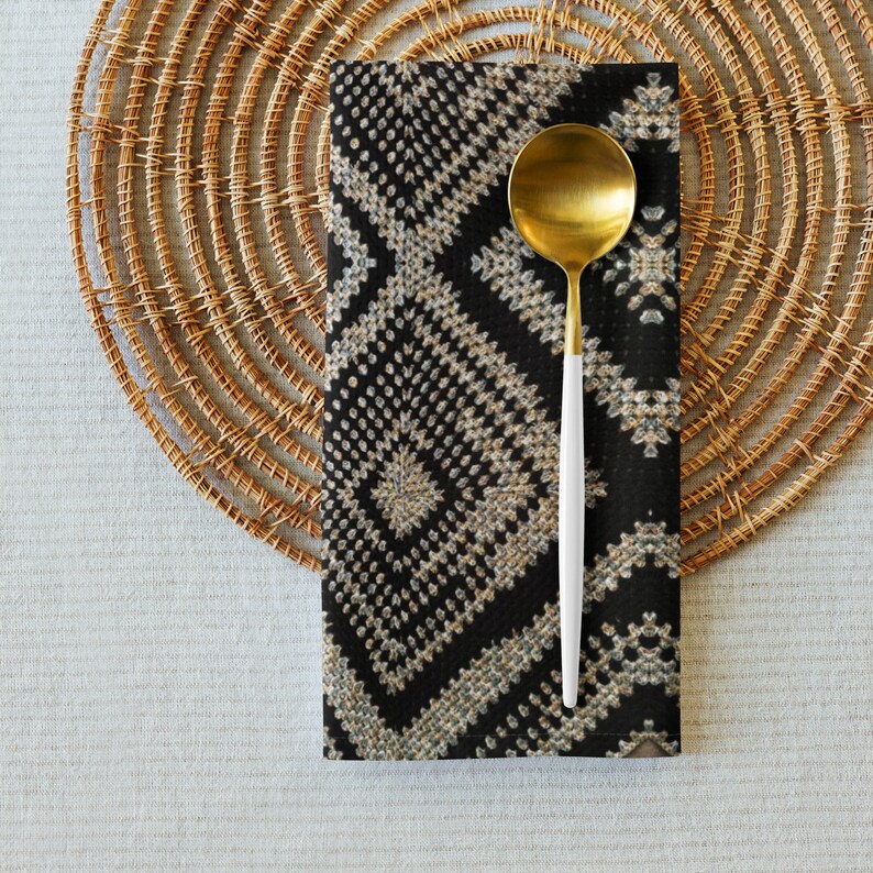 May include: A gold-colored spoon with a white handle rests on a black and beige patterned napkin. The napkin is placed on a round, woven placemat. The setting is on a light gray surface, creating a neutral dining scene.
