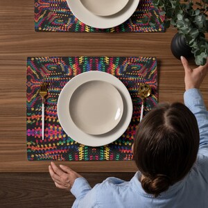 May include: Two placemats with a colorful, geometric design in shades of red, yellow, green, and blue on a black background. Each placemat has a white plate on top. A gold fork and spoon are on the placemats.