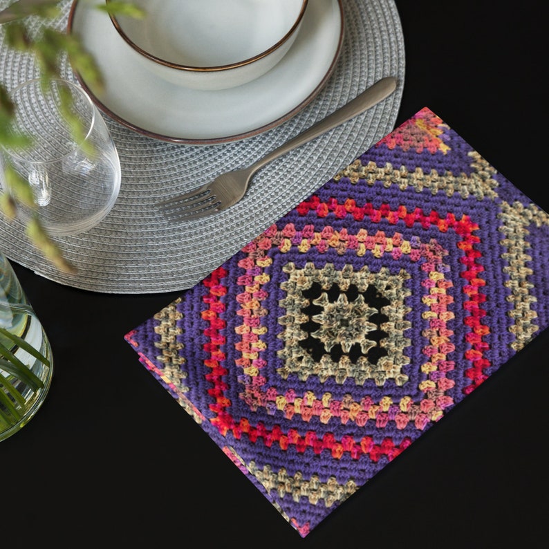 以下が含まれることがあります： グラニー・スクエア模様の正方形のかぎ針編みプレースマット。プレースマットは、ベージュの中央のデザインが特徴で、ピンク、オレンジ、紫のリングで囲まれています。白いボウル、フォーク、グラスが灰色のプレースマットの上にあります。