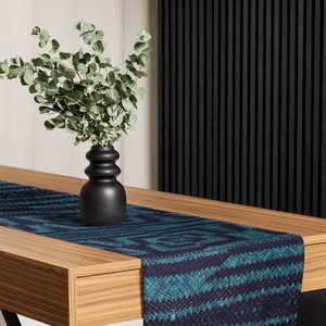 Puede incluir: Una mesa de madera con un camino de mesa estampado en azul verdoso y azul marino. Un jarr&oacute;n negro contiene un ramo de ramas de eucalipto verdes. La mesa est&aacute; colocada contra una pared de lamas negra y una pared blanca.