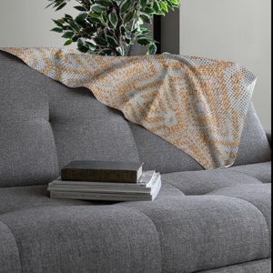 Pode incluir: Uma manta dobrada com um padr&atilde;o geom&eacute;trico em tons de bege e azul claro repousa sobre um sof&aacute; cinza. O design da manta apresenta um padr&atilde;o de pontos repetidos, criando uma apar&ecirc;ncia texturizada. Uma pilha de livros est&aacute; no sof&aacute;.