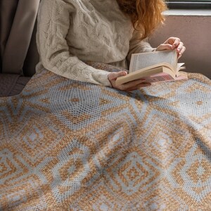 Pode incluir: Um cobertor azul claro e castanho com um padr&atilde;o geom&eacute;trico. O cobertor est&aacute; sobre uma pessoa, que est&aacute; vestindo um su&eacute;ter creme e lendo um livro. O padr&atilde;o do cobertor apresenta formas de diamante e pontos.