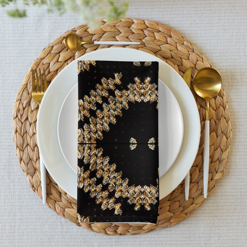 Puede incluir: Una servilleta negra doblada con un patr&oacute;n floral dorado y blanco repetido, colocada sobre un plato blanco con cubiertos dorados. El plato est&aacute; sobre un salvamanteles tejido, creando una elegante mesa.