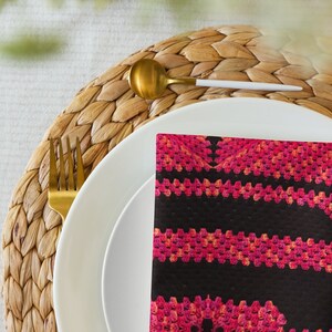 May include: A dining table setting with a white plate, gold cutlery, and a black napkin. The napkin has a pink and orange crochet design. The plate rests on a woven placemat. The scene is illuminated by soft lighting.