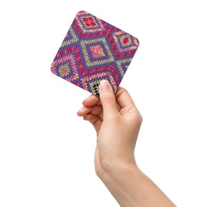 May include: A square coaster with a geometric pattern in shades of purple, pink, red, and yellow. The design features a repeating diamond pattern. The coaster is held by a hand against a white background.
