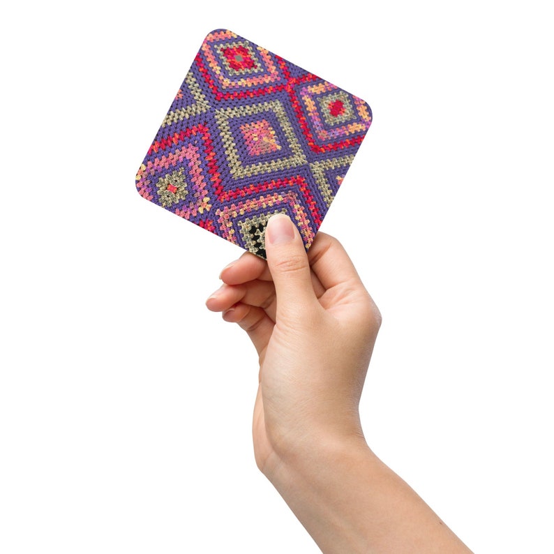 May include: A square coaster with a geometric pattern in shades of purple, pink, red, and yellow. The design features a repeating diamond pattern. The coaster is held by a hand against a white background.