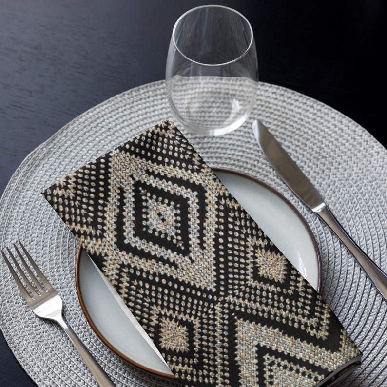 May include: A folded black and beige patterned napkin rests on a white plate, next to a knife and a wine glass. The napkin features a geometric diamond design. The plate is set on a gray placemat, with a fork to the left.