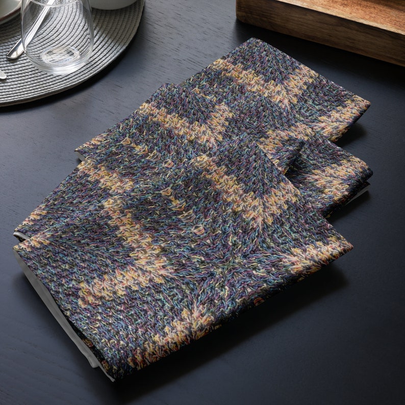 Puede incluir: Tres servilletas de tela dobladas con un patr&oacute;n de punto en tonos azules, amarillos y morados. Las servilletas est&aacute;n apiladas sobre una superficie oscura, con parte de una mesa a la vista en el fondo.