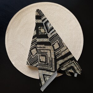 May include: A black and beige patterned napkin folded on a round, beige woven placemat. The napkin features a geometric design with squares and lines. The placemat is made of woven material and is circular in shape. The background is a dark surface.
