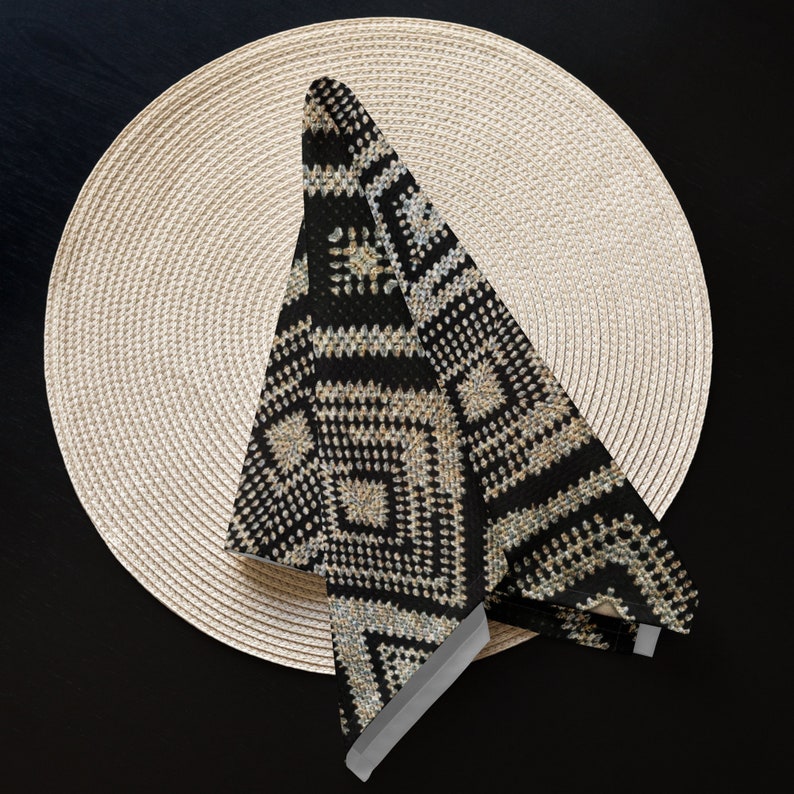 May include: A black and beige patterned napkin folded on a round, beige woven placemat. The napkin features a geometric design with squares and lines. The placemat is made of woven material and is circular in shape. The background is a dark surface.