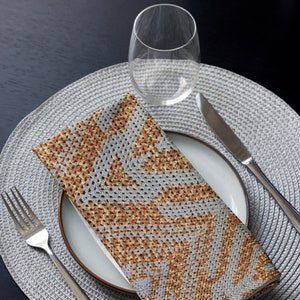 May include: A folded napkin with a grey and brown geometric pattern rests on a white plate with a brown rim. A fork and knife are placed on either side of the plate. A clear wine glass sits above the plate on a grey placemat.