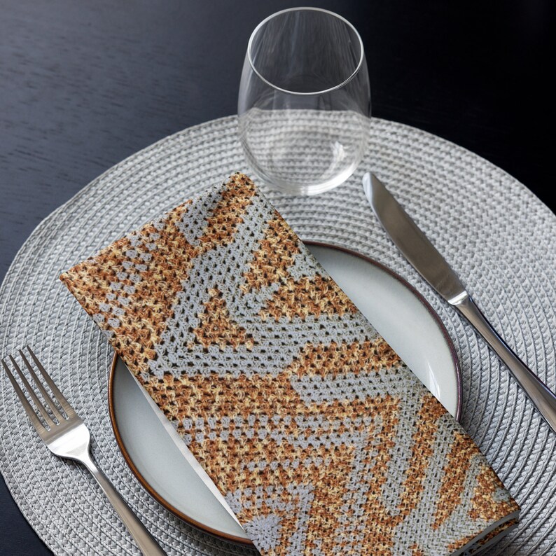 May include: A folded napkin with a grey and brown geometric pattern rests on a white plate with a brown rim. A fork and knife are placed on either side of the plate. A clear wine glass sits above the plate on a grey placemat.
