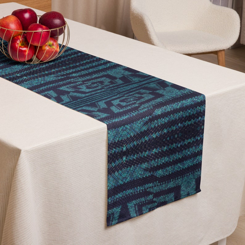 Puede incluir: Un camino de mesa con estampado en azul verdoso y azul marino. El camino de mesa presenta un dise&ntilde;o geom&eacute;trico con l&iacute;neas horizontales y verticales. El camino de mesa se muestra sobre un mantel blanco, con una cesta de alambre dorada con manzanas rojas en el fondo.