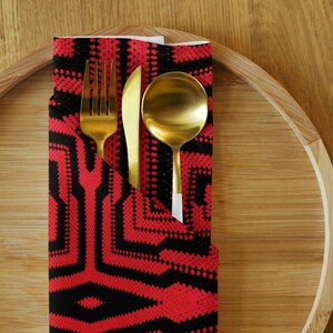 May include: A gold-colored fork, knife, and spoon set rests in a red and black patterned napkin. The napkin is folded and placed on a bamboo tray. The utensils are arranged for a formal dining setting.