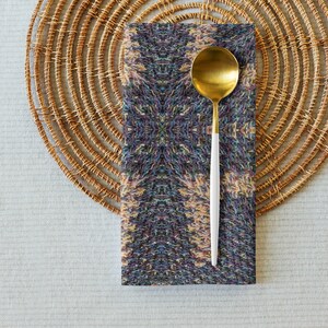 Puede incluir: Una cuchara dorada con mango blanco descansa sobre una servilleta estampada. La servilleta presenta un dise&ntilde;o sim&eacute;trico con una mezcla de colores, incluyendo azul, morado y amarillo. La servilleta est&aacute; colocada sobre un salvamanteles circular tejido.