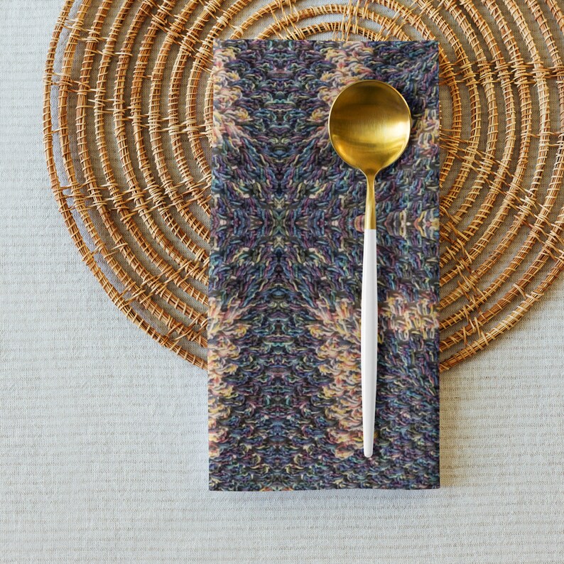 Puede incluir: Una cuchara dorada con mango blanco descansa sobre una servilleta estampada. La servilleta presenta un dise&ntilde;o sim&eacute;trico con una mezcla de colores, incluyendo azul, morado y amarillo. La servilleta est&aacute; colocada sobre un salvamanteles circular tejido.