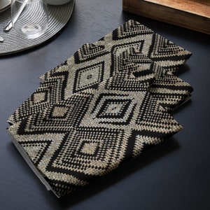May include: Four folded black and beige geometric patterned cloth napkins. The napkins feature a repeating diamond design. The napkins are made of a textured fabric and are stacked on top of each other. The napkins are on a dark surface.