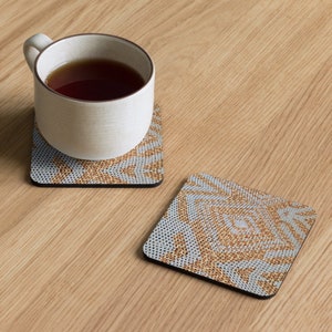 May include: Two square coasters with a geometric pattern in gold and pale blue, one with a white mug filled with tea. The coasters have a black border and are on a light wood surface.