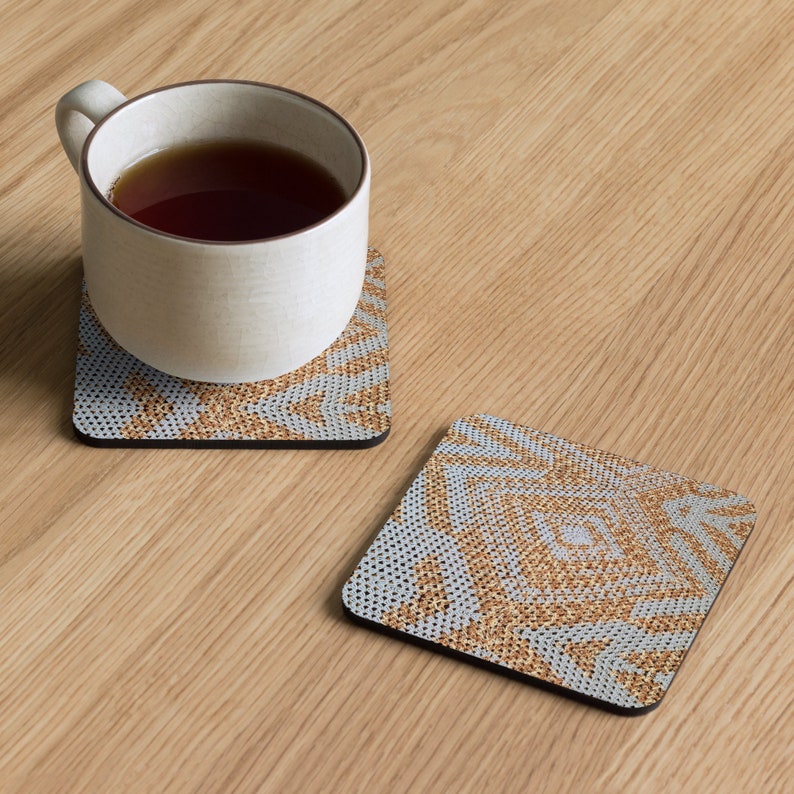 May include: Two square coasters with a geometric pattern in gold and pale blue, one with a white mug filled with tea. The coasters have a black border and are on a light wood surface.