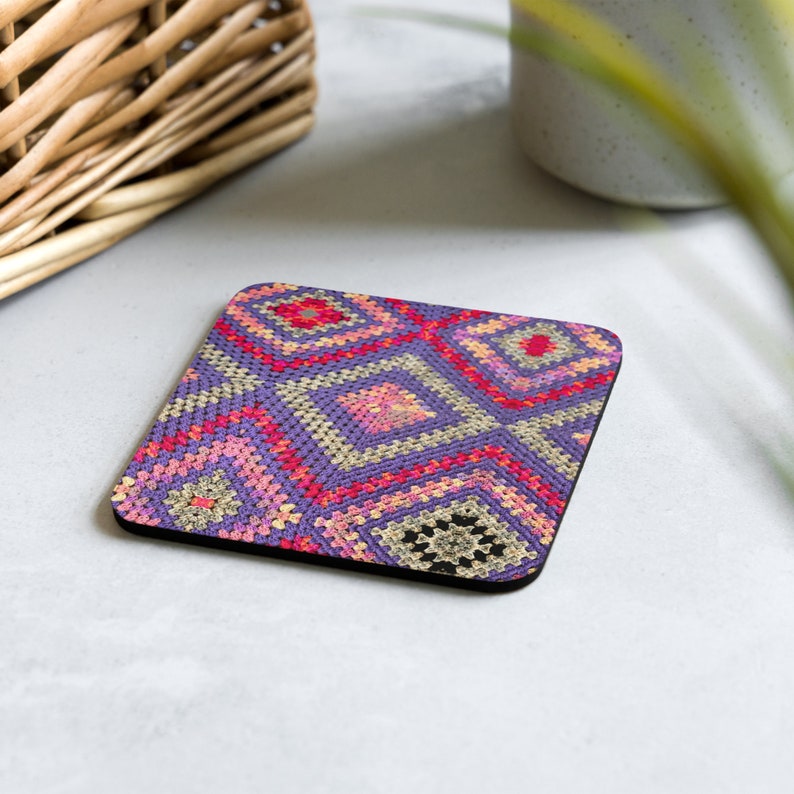 May include: Square coaster with a colorful crochet pattern in shades of purple, pink, red, and beige. The coaster has a black border and a geometric design, perfect for protecting surfaces from drink rings.