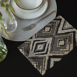 May include: A black and beige geometric patterned placemat is placed on a black surface. A gray woven placemat, a white bowl with a brown rim, and a silver fork are arranged on top. A clear glass and a vase with green stems are also visible.