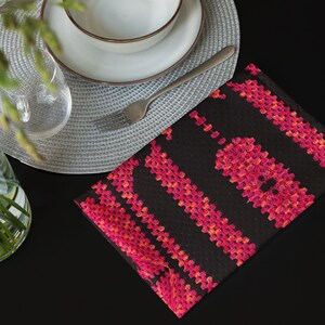 May include: A black napkin featuring a repeating pattern of pink and orange floral designs. The napkin is placed on a black surface, next to a place setting with a white plate, bowl, fork, and a glass.