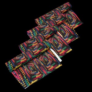 Op de afbeelding: Zwarte stof met een kleurrijk, geometrisch patroon in rood, geel, groen en blauw. De stof is opgerold en gevouwen, waardoor het ontwerp wordt getoond. Het patroon lijkt op een haak- of breistijl.