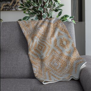 Pode incluir: Uma manta dobrada com um padr&atilde;o geom&eacute;trico em tons de bege e cinza claro repousa sobre um sof&aacute; cinza. O design da manta apresenta formas de diamante e espiral. As bordas s&atilde;o finalizadas com uma borda branca, adicionando um contraste limpo.