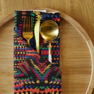 Op de afbeelding: Een gouden vork, mes en lepel set in een zwarte servet met een kleurrijk, geometrisch patroon. De servet ligt op een bamboe serveerblad, wat zorgt voor een stijlvolle tafelsetting.