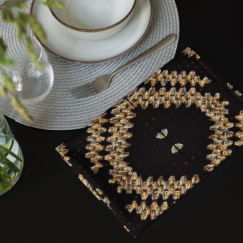 Puede incluir: Una servilleta cuadrada negra con un patr&oacute;n repetido de formas abstractas doradas y marrones. La servilleta est&aacute; doblada y colocada sobre una superficie negra, junto a un salvamanteles gris, un tenedor y un cuenco.