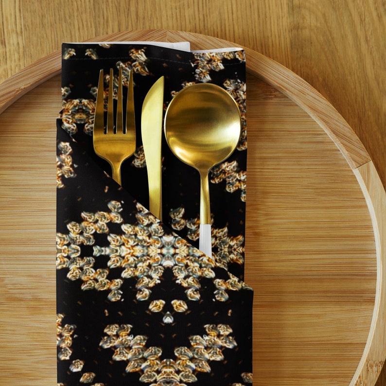 Puede incluir: Un juego de tenedor, cuchillo y cuchara dorados sobre una servilleta negra con un estampado floral. La servilleta est&aacute; doblada y colocada sobre una bandeja de madera, creando una elegante mesa. Los utensilios son de metal.