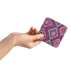May include: A square coaster with a geometric pattern in shades of purple, red, pink, and yellow. The coaster is held in a hand against a white background. The design features diamond shapes.