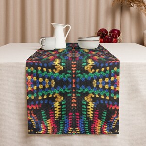 May include: A black table runner with a vibrant, geometric pattern in red, yellow, blue, and green. The runner is placed on a white tablecloth, with a white pitcher, cup, and bowls visible in the background. The runner adds a pop of color.