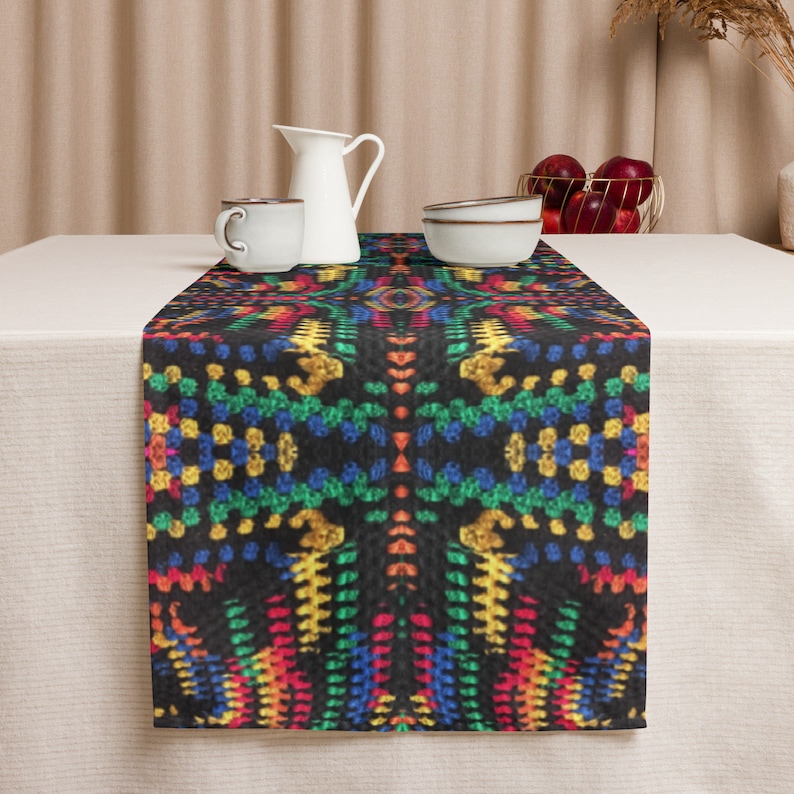 May include: A black table runner with a vibrant, geometric pattern in red, yellow, blue, and green. The runner is placed on a white tablecloth, with a white pitcher, cup, and bowls visible in the background. The runner adds a pop of color.