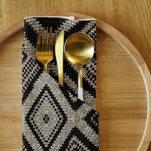 May include: A gold-colored fork, knife, and spoon set rests in a black and white patterned napkin. The napkin is folded and placed on a bamboo serving tray. The utensils have a shiny finish.