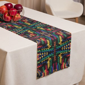 May include: A black table runner with a vibrant, multicolored pattern of red, yellow, green, and blue. The runner is placed on a white tablecloth, with a gold wire basket of red apples in the background.