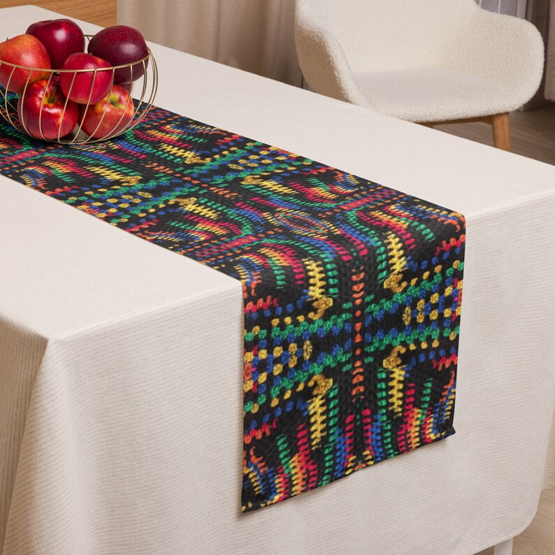 May include: A black table runner with a vibrant, multicolored pattern of red, yellow, green, and blue. The runner is placed on a white tablecloth, with a gold wire basket of red apples in the background.