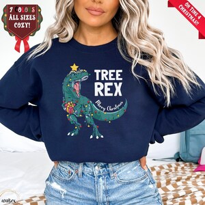 Könnte beinhalten: Marineblaues Sweatshirt mit einem grünen Dinosaurier, der mit Weihnachtsbeleuchtung und einem Stern geschmückt ist. Der Text "TREE REX Merry Christmas" ist weiß. Ein rotes Banner zeigt "Oh Time 4 Christmas!!".