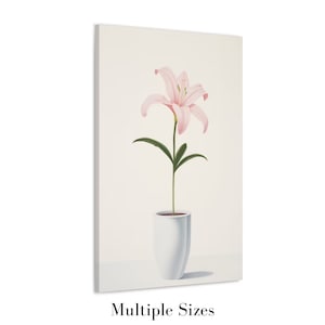 以下が含まれることがあります： 白い花瓶に入ったピンク色のユリのキャンバスプリント。花は繊細な花びらと緑の葉を持っています。花瓶はシンプルでモダンなデザインです。「Multiple Sizes」の文字が下にあります。