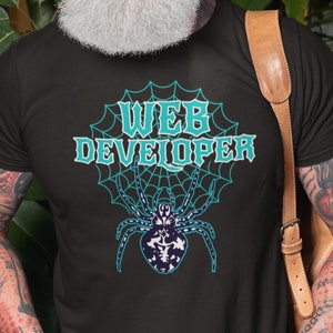 May include: Black t-shirt with the words "WEB DEVELOPER" in teal and a teal spider web design. The spider in the center is white and navy blue. The person wearing the shirt is smiling.